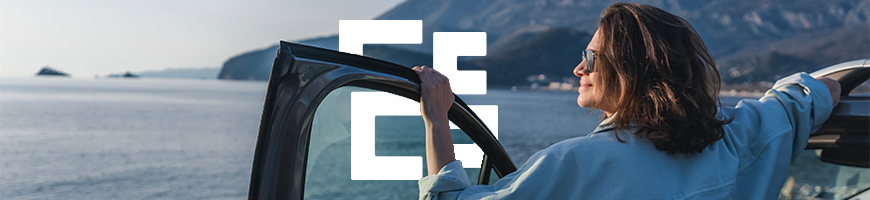 Woman stands by open car door smiling at the view of a body of water with mountains in the background