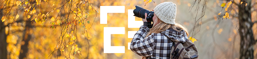 A woman wearing a black and white gingham jacket and a cream beanie take photos of an autumnal forest with a large black DSLR camera.