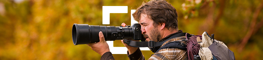 A man wearing a gingham shirt and large backpack takes photos using a long lens camera in an autumnal landscape.