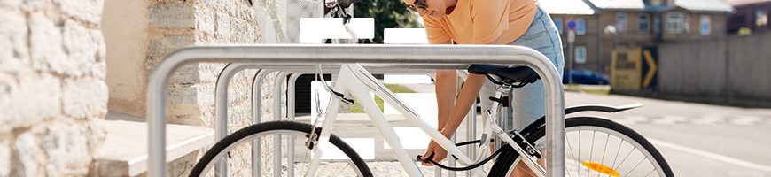 A woman locks a white bicycle to a metal bike rack on a sunny street.