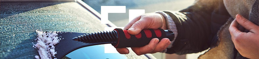 A person wearing a scarf scrapes frost off a car windshield with a red and black ice scraper.