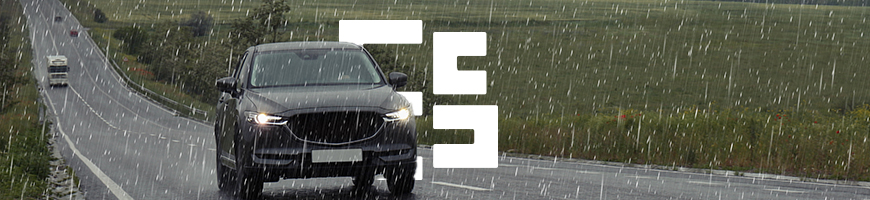A black car drives on a wet rural road during heavy rain with stormy clouds overhead and fields alongside.
