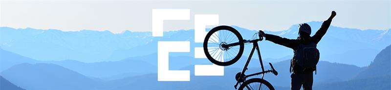 Person stood on top of a cliff holding a bike in the air in celebration with mountains in the background