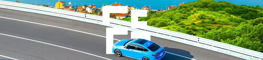 A blue car drives on a curved coastal road with lush green hills to the right and a serene blue sea in the background