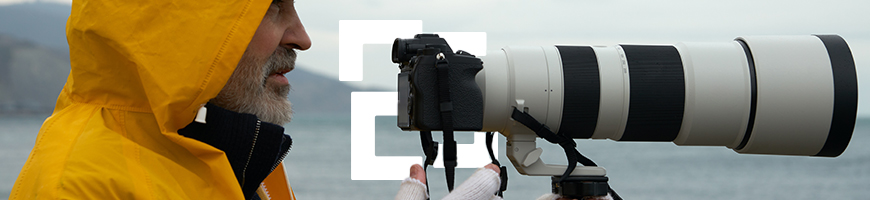 Man in a yellow raincoat with his hood up under an overcast sky holds a large camera with a telephoto lens by the sea
