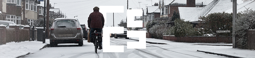 A person in a jacket cycles down a snow-covered residential street lined with houses and parked cars.