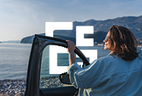 Woman stands by open car door smiling at the view of a body of water with mountains in the background