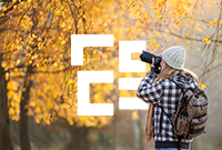 A woman wearing a black and white gingham jacket and a cream beanie take photos of an autumnal forest with a large black DSLR camera.