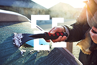 A person wearing a scarf scrapes frost off a car windshield with a red and black ice scraper.