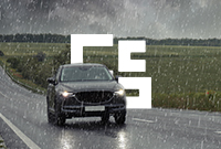 A black car drives on a wet rural road during heavy rain with stormy clouds overhead and fields alongside.