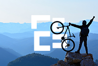 Person stood on top of a cliff holding a bike in the air in celebration with mountains in the background