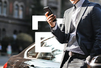 Man wearing a suit leans against a black car looking at his smartphone