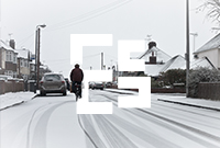 A person in a jacket cycles down a snow-covered residential street lined with houses and parked cars.