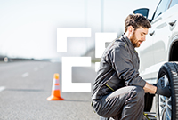 A man changes a tyre on a car parked on the roadside with an orange safety cone placed behind the vehicle